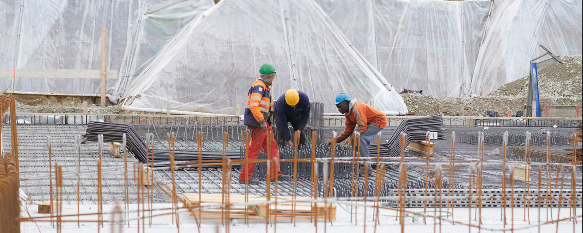 건설 현장에서 작업자들이 철근을 다루고 있으며, 뒤에는 비닐로 덮인 구조물이 보입니다.